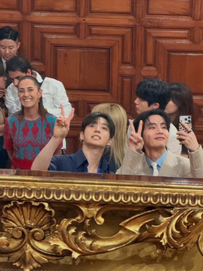 A group of people smiling and posing for a photo in front of a decorative wooden backdrop. Two individuals in the foreground are making peace signs, while one is taking a selfie. There are other attendees visible in the background.