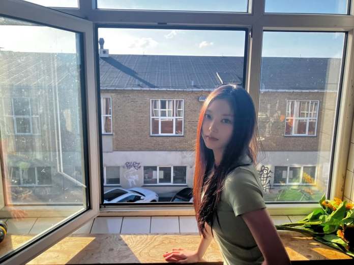 Haerin with long dark hair sitting by a window, looking towards the camera, with sunlight filtering in and buildings visible outside.