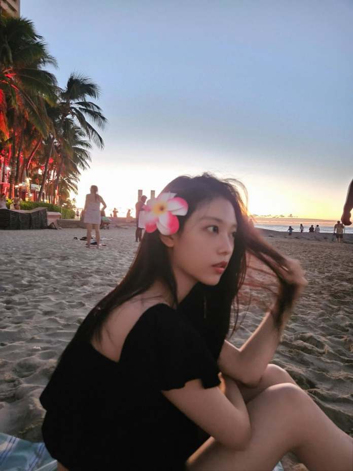 Haerin with a flower in her hair sitting on the beach at sunset, looking pensive as she brushes her hair back. Palm trees can be seen in the background.