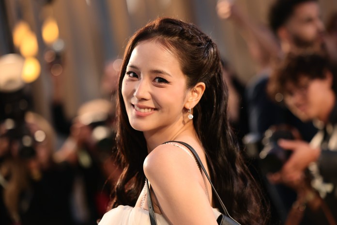 Jisoo with long dark hair, wearing a stylish dress and earrings, poses for the camera at an event, with photographers and a crowd in the background.