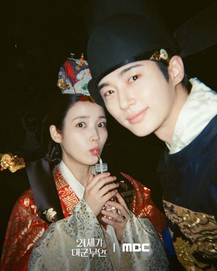 IU in traditional Korean clothing holding a drink with a straw, and Byeon Woo-seok in historical attire smiling beside her, set against a dark background.