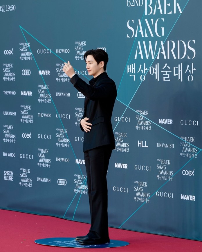 Lee Junho in a black suit poses confidently on the red carpet at the 62nd Baek Sang Awards, with a backdrop featuring event logos and sponsors.
