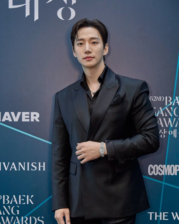 Lee Junho wearing a black suit and shirt poses confidently in front of a blue backdrop featuring event logos.