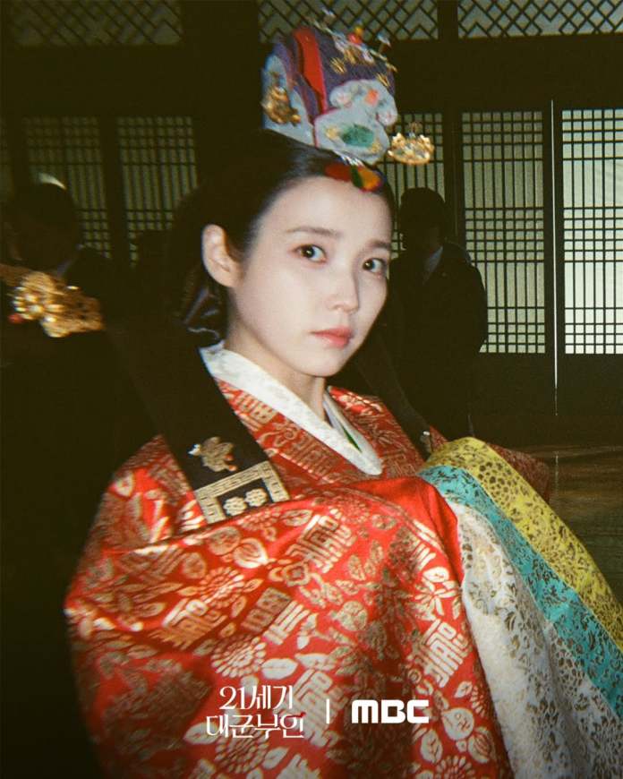 A close-up portrait of IU in traditional Korean hanbok, featuring a vibrant red and patterned garment and an ornate hairpiece, set against a historical backdrop.