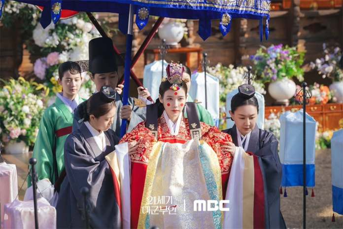 A traditional Korean ceremony featuring IU in ceremonial attire surrounded by attendants in historical clothing, with decorative flowers and a festive ambiance.