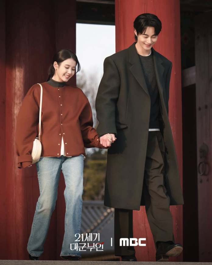 A smiling IU in a brown oversized jacket and blue jeans holds hands with Byeon Woo-seok in a black coat as they walk together in an outdoor setting with traditional architecture.