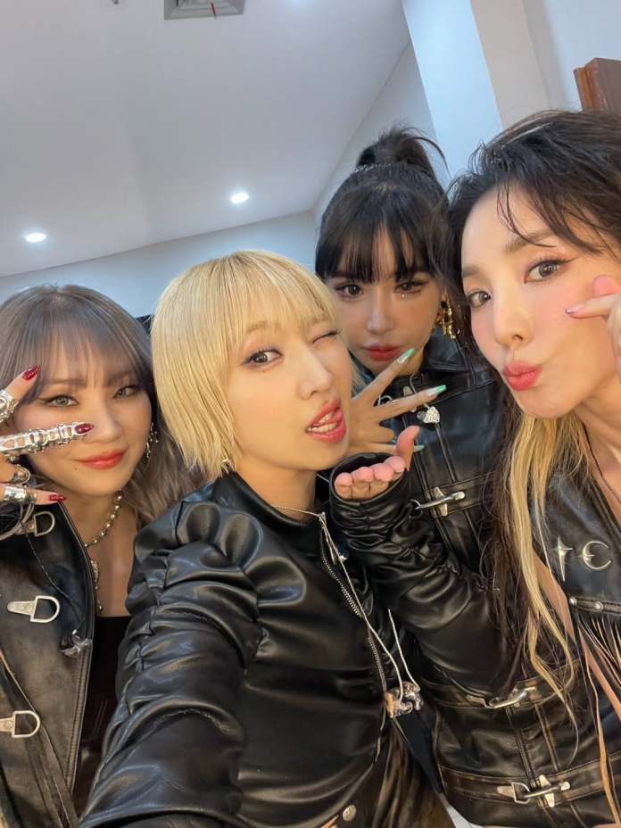 2ne1 members posing for a selfie, wearing stylish black leather jackets and showing playful expressions in a brightly lit room.
