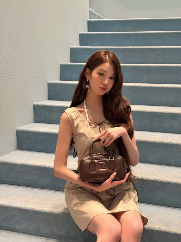 Jang Wonyoung sitting on blue carpeted steps, wearing a light beige dress, holding a small brown handbag, with long, wavy hair and earrings.
