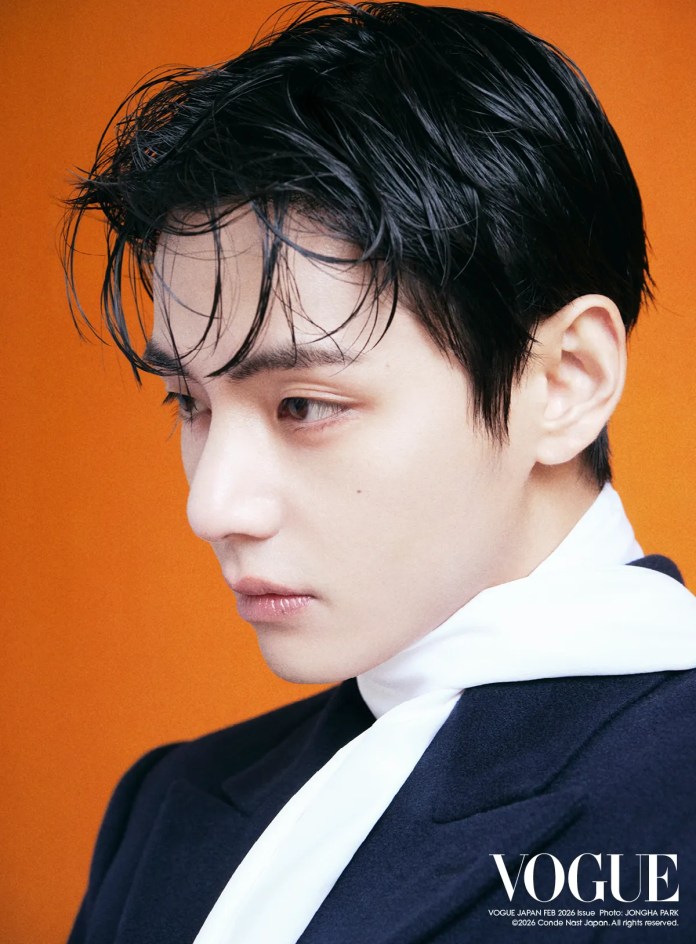 Profile shot of a young man with sleek, dark hair, wearing a black jacket and white scarf against an orange background.