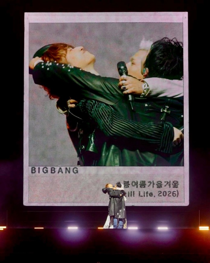 A performance scene featuring members of Big Bang embracing on stage, with a large graphic display of a photograph behind them, labeled 'BIGBANG'.