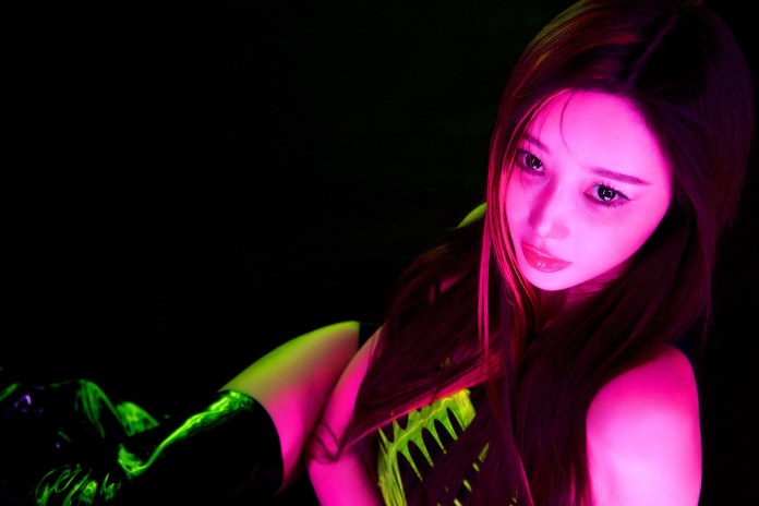 A close-up portrait of a person with long hair, styled in a fashionable manner, seated against a dark background with vibrant neon lighting casting pink and green hues on their face.