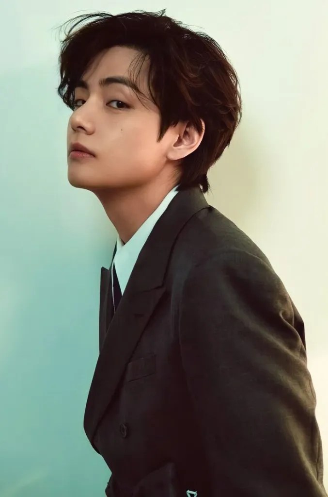 A person in a stylish suit with dark hair, posing to the side against a light background.