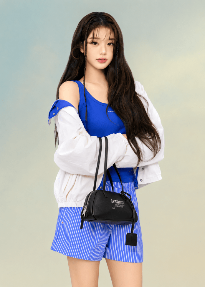 Jang Wonyoung with long hair wearing a blue tank top and striped shorts, holding a black handbag. She stands against a softly colored background, exuding a casual and stylish vibe.