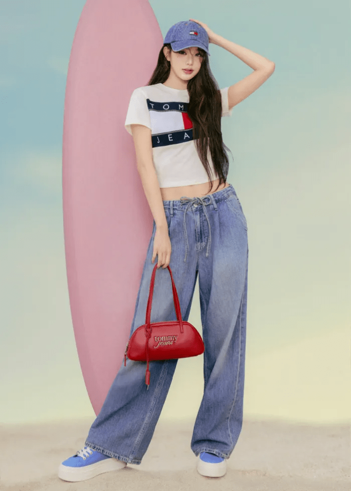 Jang Wonyoung stands confidently beside a pink surfboard, wearing a white cropped T-shirt with a blue and red logo, loose denim jeans, and blue sneakers. She holds a small red handbag and wears a matching blue cap.