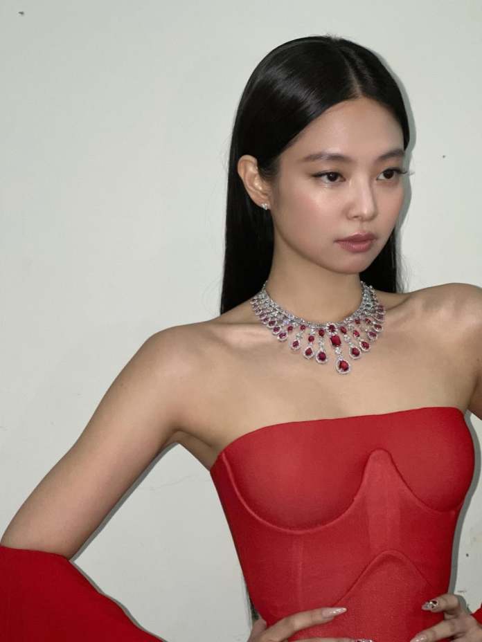  JENNIE wearing a strapless red dress and an elegant necklace, posing confidently against a neutral background.