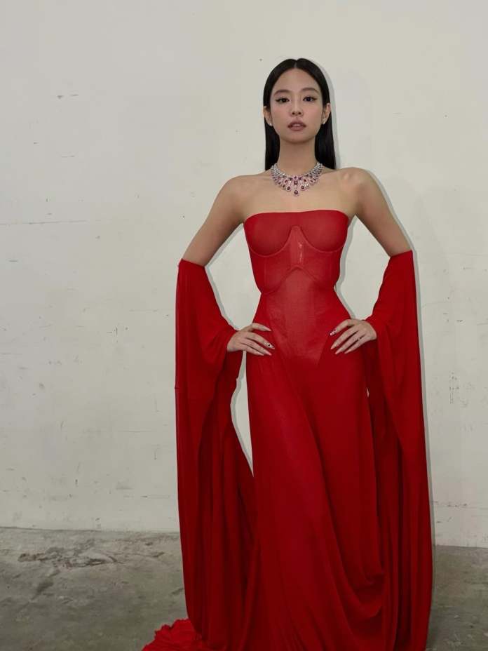 JENNIE in a striking red, floor-length dress with long flowing sleeves, posing confidently against a neutral background.