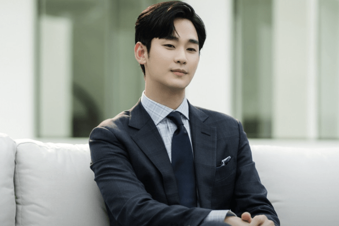 Kim Soo-hyun in a tailored suit sitting on a couch, with a calm expression, against a modern interior backdrop.