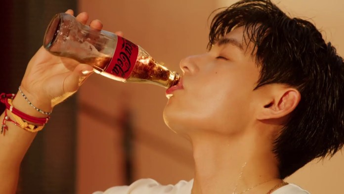 V with short dark hair drinks from a glass Coca-Cola bottle, tilting his head back as he savors the beverage.