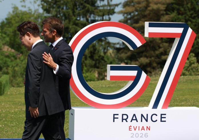 Presidents of South Korea and France walk past a large 'G7' sign, featuring the colors of the French flag, set in a green outdoor area during a summit.