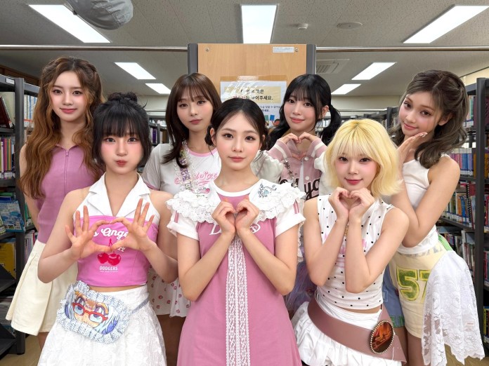 UNIS posing together in a library, all wearing pink outfits and making heart shapes with their hands.