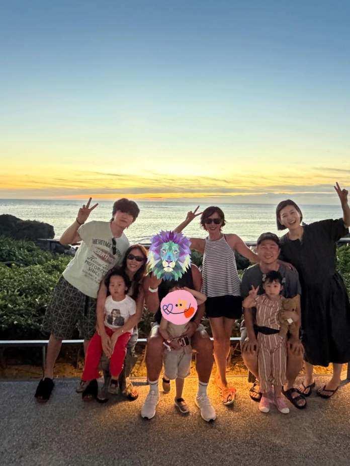 A group of seven people posing for a photo at sunset by the ocean. They are smiling and showing peace signs, with children sitting in front.
