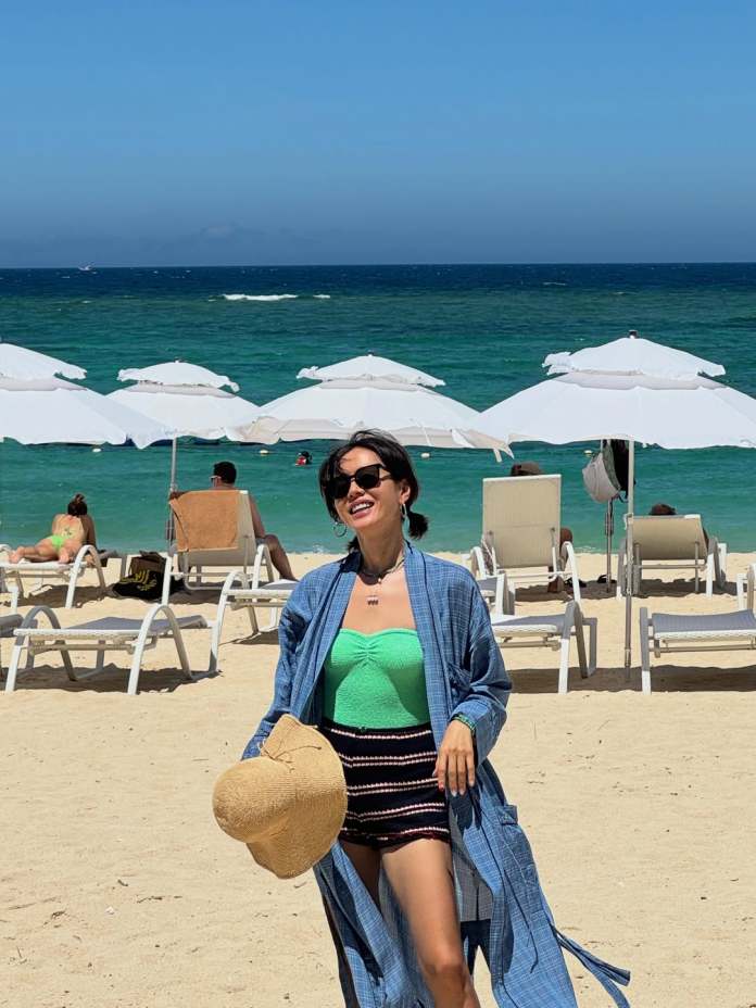 Son Ye Jin in a green top and striped shorts smiles while walking on the beach, holding a straw hat. Beach umbrellas and sun loungers are in the background with a turquoise ocean and clear blue sky.