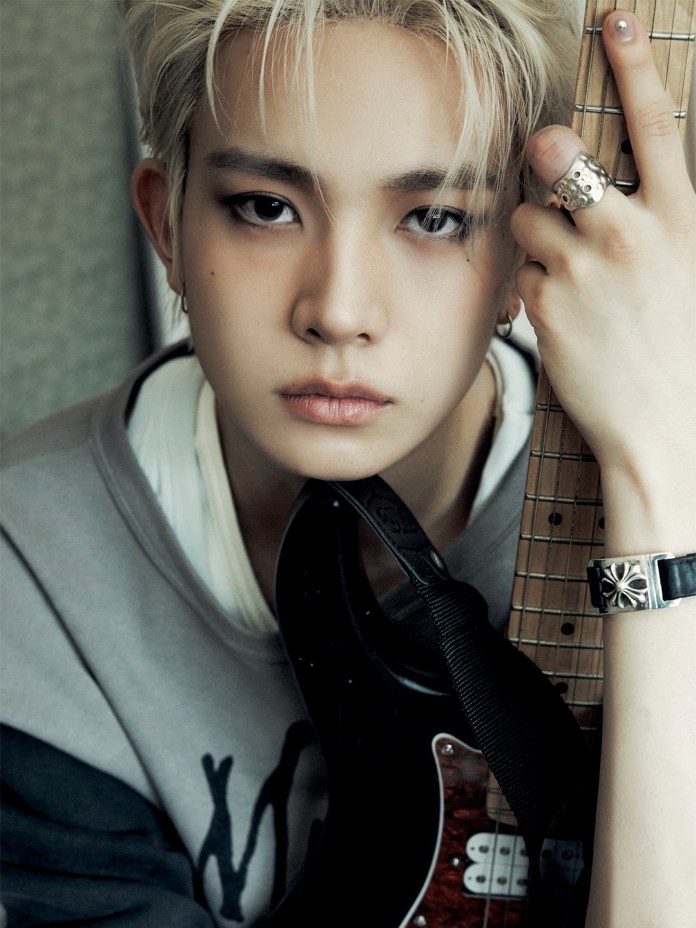 Close-up portrait of EVAN with blonde hair, wearing a gray sweatshirt and holding an electric guitar. The individual has striking eye makeup and multiple rings, with a piercing in one ear.