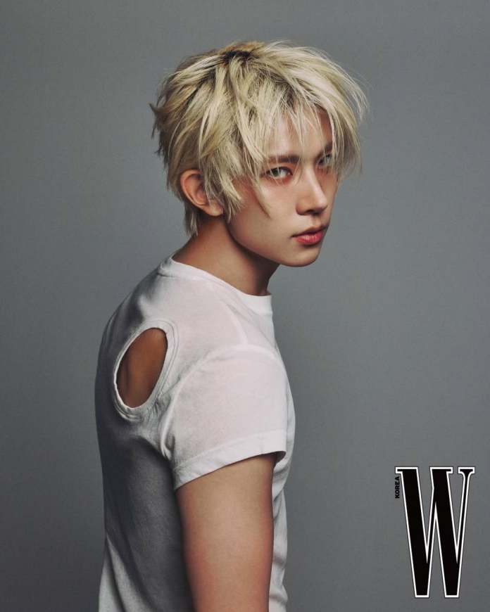 EVAN with tousled blonde hair is posing sideways, wearing a fitted white t-shirt with a cutout on one shoulder. The background is a soft gray, creating a minimalist look.
