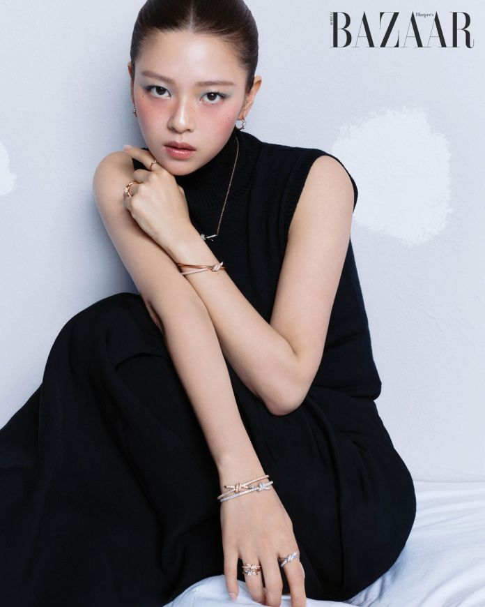 Jeongyeon wearing a black sleeveless top and a flowing skirt, posing with arms crossed and showcasing various pieces of jewelry, including rings and bracelets. The background features a light gray wall.