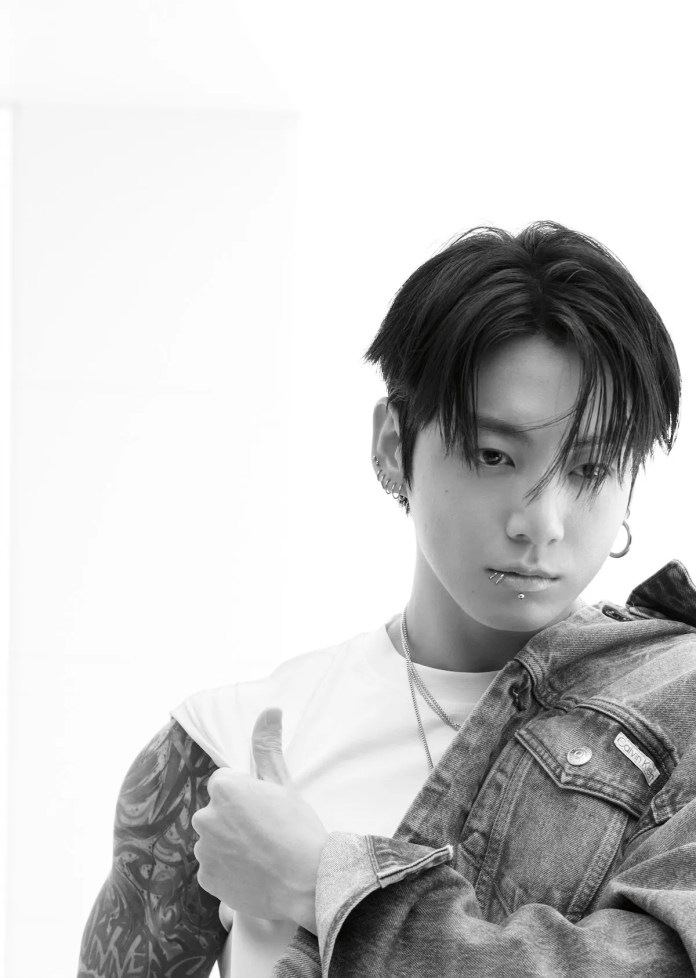 Jungkook with dark hair styled in a casual look, wearing a white t-shirt and a denim jacket. He is giving a thumbs up with his left hand and has a visible tattoo on his right arm. The image is in black and white with a minimalistic background.