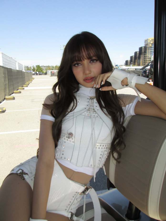Lisa wearing a stylish white outfit with accessories, sitting on a vehicle in an outdoor setting with buildings and a clear blue sky in the background.