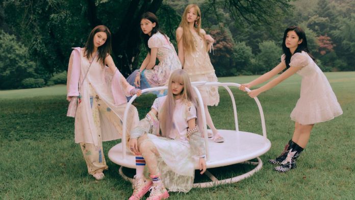 A group of five young women in pastel-colored dresses posing on a circular platform in a grassy outdoor setting, surrounded by trees.