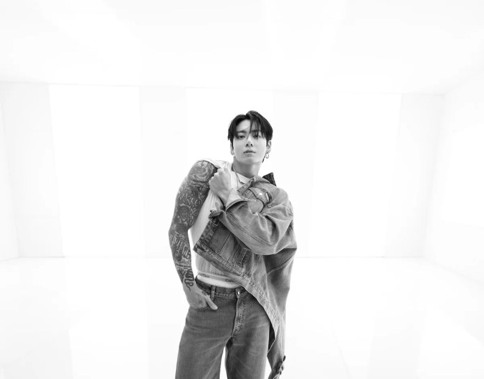Jungkook poses confidently in a minimalist, brightly lit setting, wearing a denim jacket and showing off intricate tattoos.