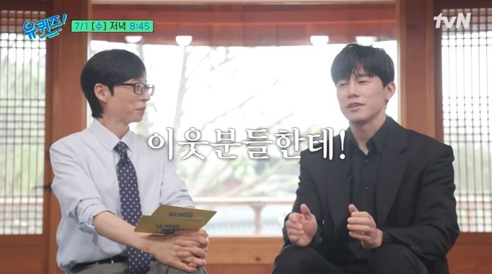 Kim Mu-yeol are seated indoors in a traditional setting, engaging in conversation. One man, wearing glasses and a light blue shirt with a tie, is listening attentively while holding a card. The other man, dressed in black, appears to be speaking animatedly. Text overlay in Korean is present in the foreground.