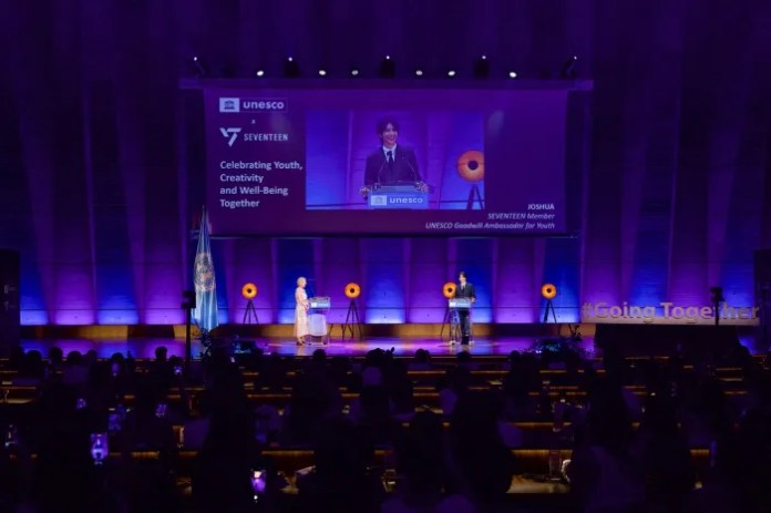 A stage event featuring a speaker at a podium with a large screen displaying event details. The audience is seated at tables, and the venue is illuminated with purple lighting. The event celebrates youth and creativity in collaboration with UNESCO and SEVENTEEN.