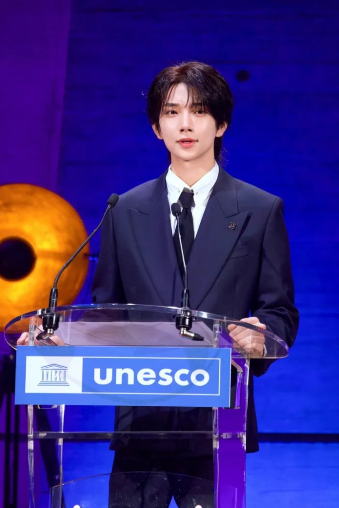 A young man in a suit stands at a podium with a UNESCO sign, speaking into a microphone under dramatic lighting.