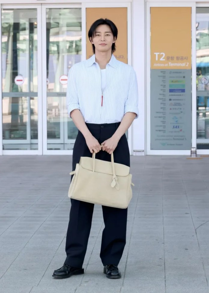 Park Seo-joon standing outside an airport terminal, wearing a striped button-up shirt and black trousers, holding a beige handbag.