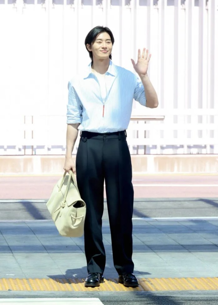 Park Seo-joon standing on a sidewalk, waving and smiling, wearing a light blue striped shirt and black trousers, holding a beige bag.