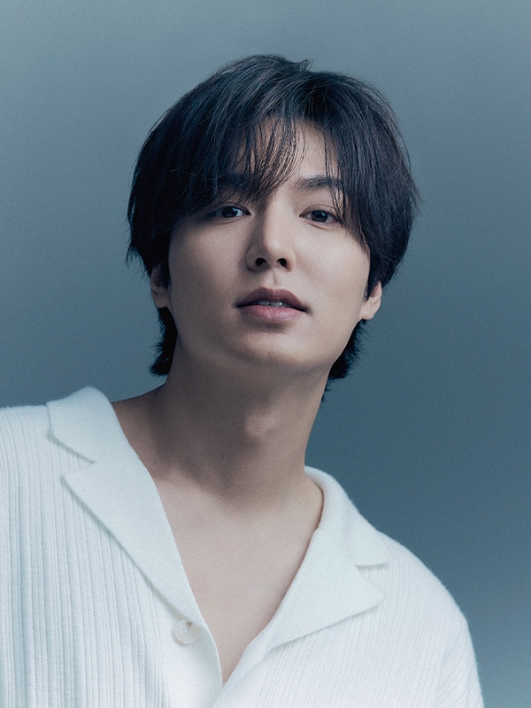 Lee Min-ho with soft features and light, tousled hair, wearing a white sweater with a collar, against a blue background.