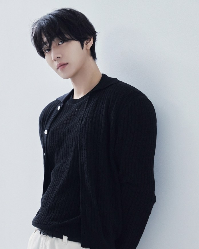 Ahn Hyo-seop with dark hair, wearing a black ribbed sweater and a cardigan, stands against a light gray wall, looking confidently at the camera.