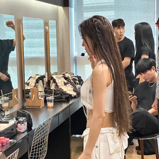 Ha Ji-won with long, braided hair stands in front of a mirror in a dressing room, preparing for a photoshoot. Several people are present in the background, including a stylist and others seated at a makeup station.