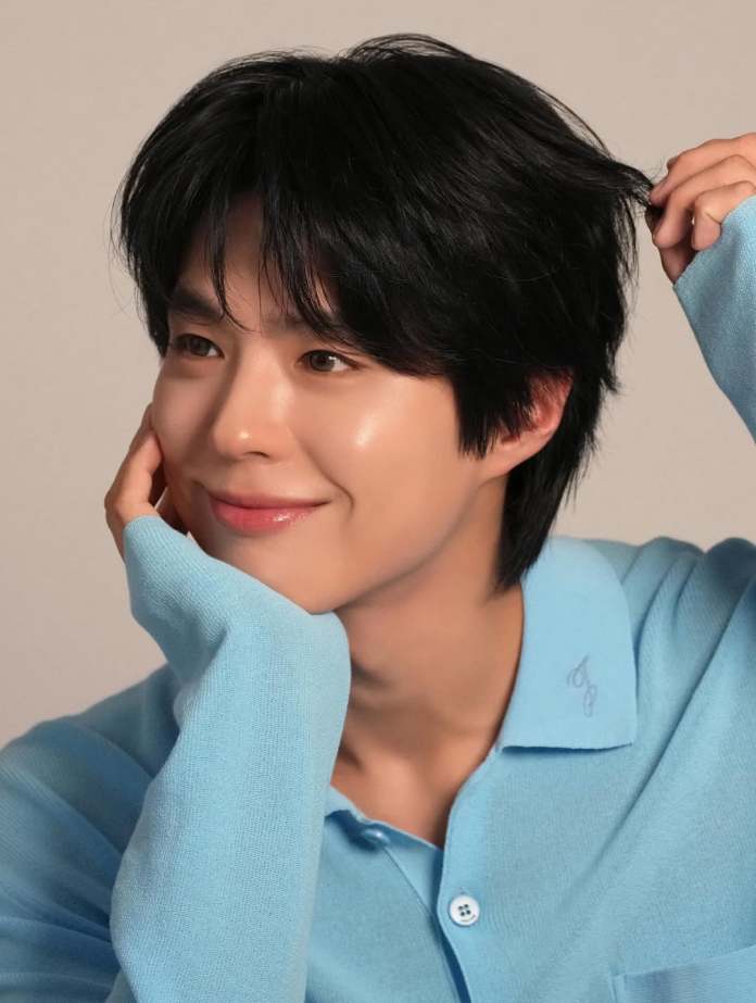 Park Bo Gum with black hair, wearing a light blue sweater, gently holds a strand of his hair while resting his chin on his hand.