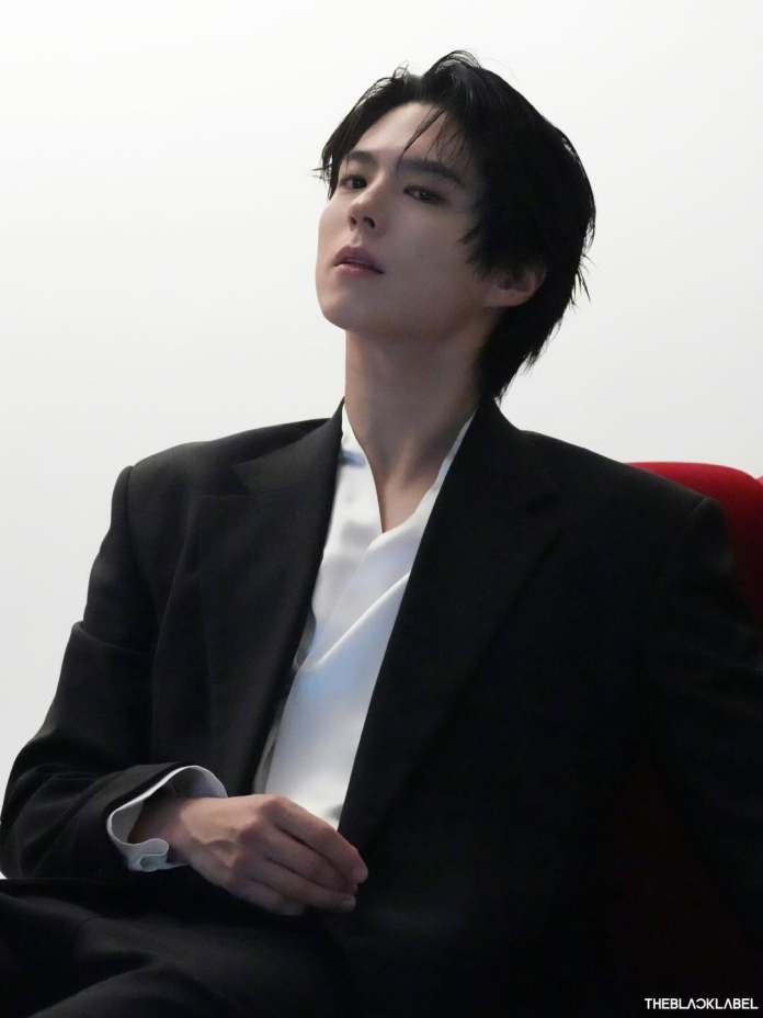 Park Bo Gum with dark, tousled hair dressed in a black suit and a white shirt, sitting in a relaxed pose with a confident expression.
