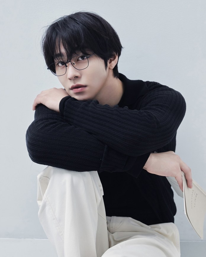Ahn Hyo-seop with black hair and glasses sitting against a light-colored wall, dressed in a black top and white pants, holding a book while looking thoughtfully at the camera.