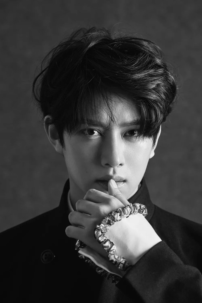 Kim Heechul with tousled hair, wearing a high-collared dark outfit, posed thoughtfully with one hand near his face.