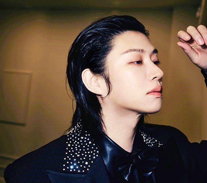 Kim Heechul with long, slicked-back hair wearing a black suit with embellished shoulders, lightly touching their hair.
