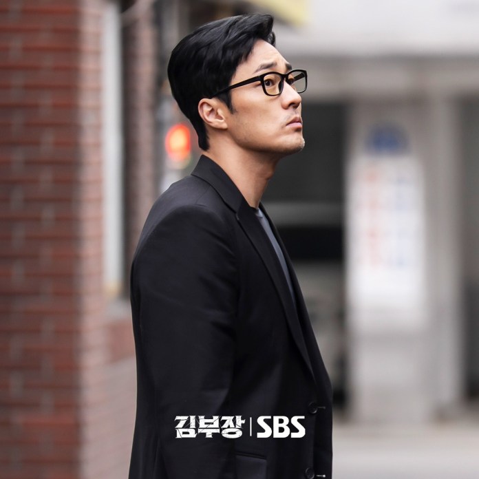So Ji-sub in a black suit and glasses stands sideways with a thoughtful expression, looking upwards in an outdoor urban setting.