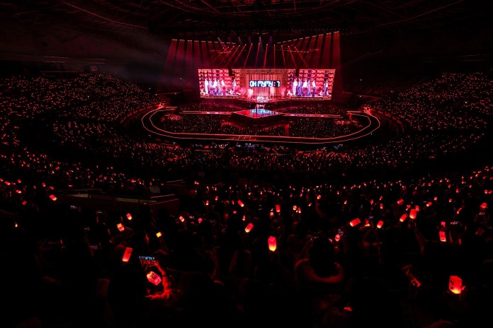 A large crowd at a concert, with many fans holding red glow sticks. The stage is brightly lit, featuring tws and a vibrant backdrop, creating a dynamic atmosphere.