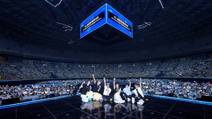 TWS on stage in a large arena, surrounded by an enthusiastic audience. The performers strike a pose while the crowd holds up lightsticks and banners, creating a vibrant atmosphere.