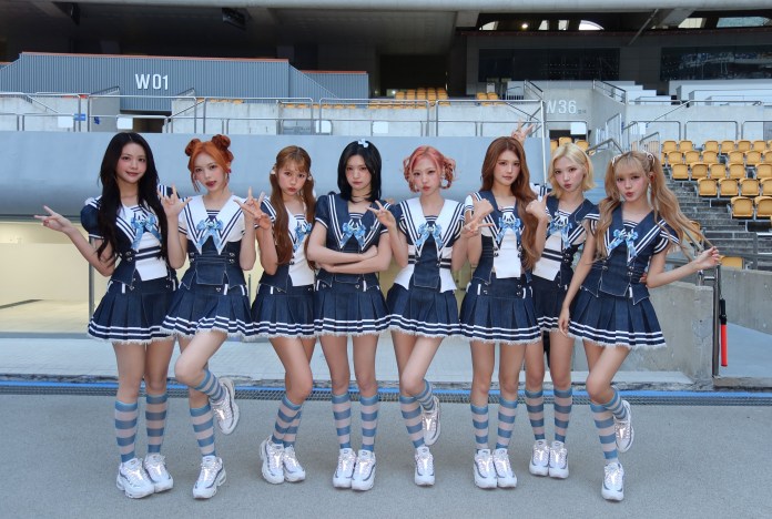 Hearts2Hearts posing in coordinated outfits featuring sailor-style tops and skirts, with striped knee-high socks, in a stadium setting.